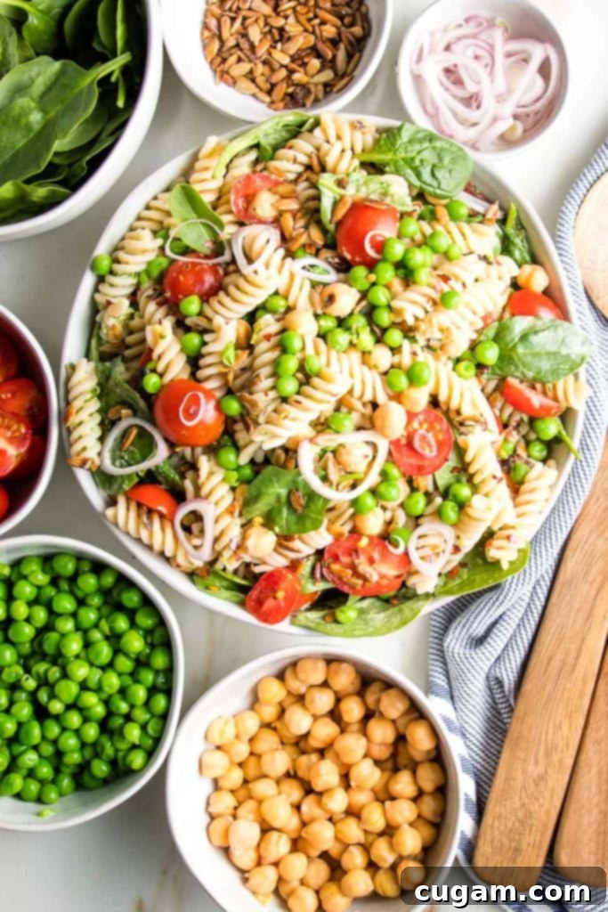 Simple Summer Pasta Salad 2 Small white bowls filled with chickpeas, green peas, halved cherry tomatoes, spinach, sunflower seeds, and sliced shallots arranged around a large white bowl of colorful pasta salad.