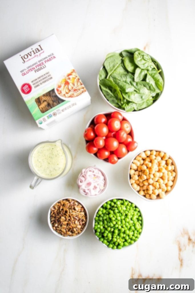 Simple Summer Pasta Salad 3 Top-down view of gluten-free fusilli pasta, a pitcher of green tahini sauce, and individual bowls containing fresh spinach, cherry tomatoes, sliced shallots, shelled sunflower seeds, green peas, and chickpeas.