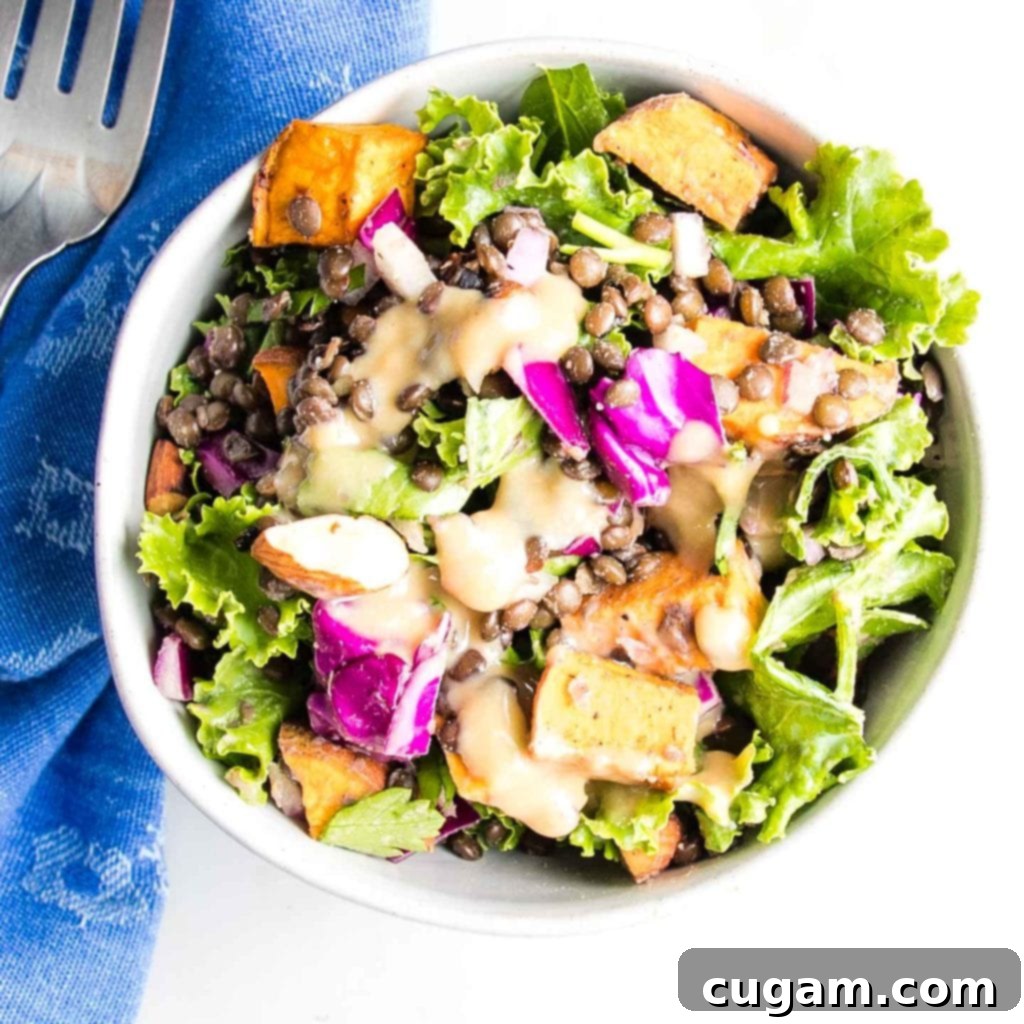 A white bowl showcasing a perfectly dressed lentil salad, featuring kale, cabbage, sweet potato cubes, black lentils, onions, almonds, parsley, and a creamy, inviting dressing.