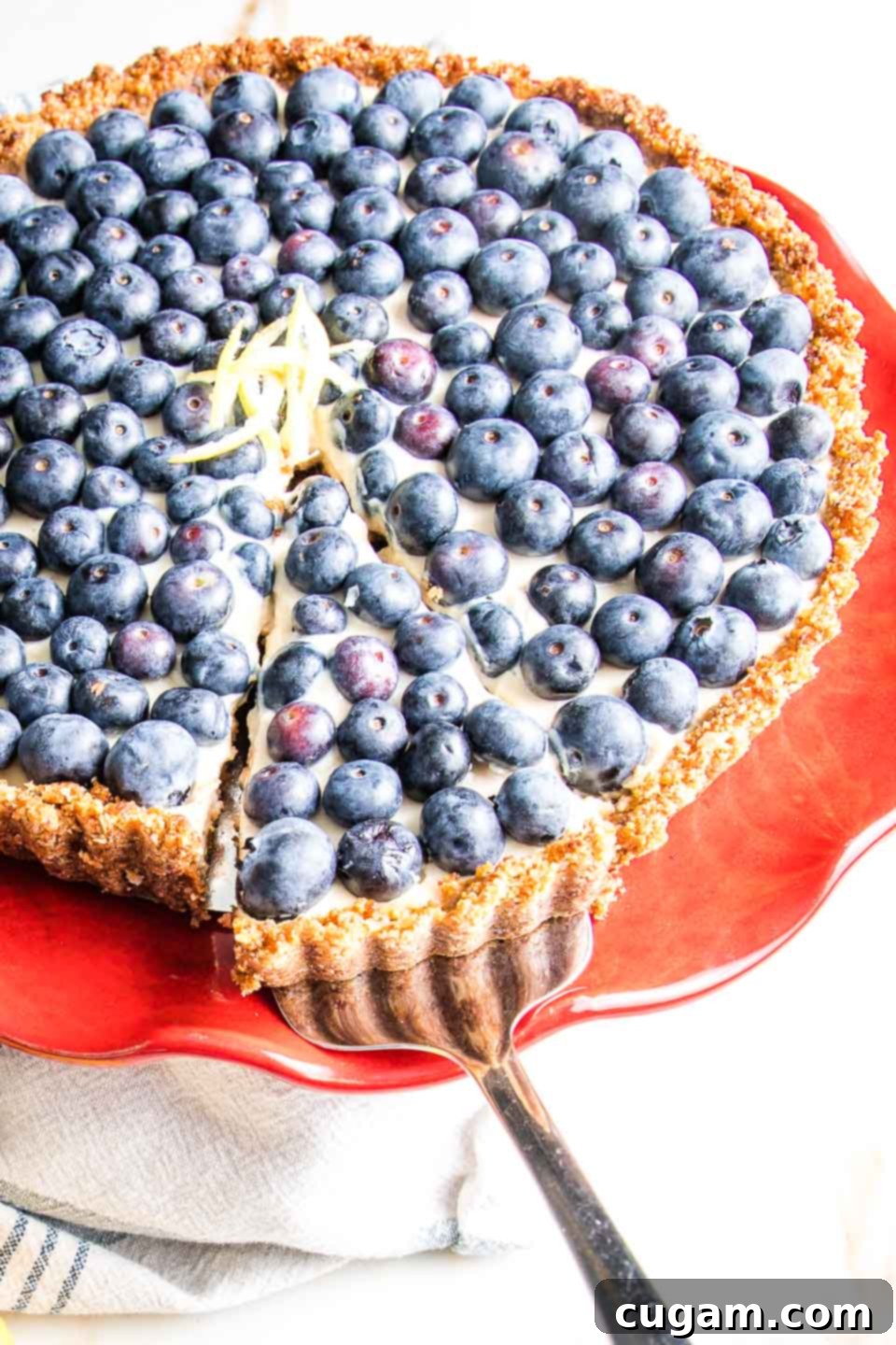 A hand gently pulling out a perfect slice of the blueberry tart, revealing its creamy layers, set against a bright red cake stand.