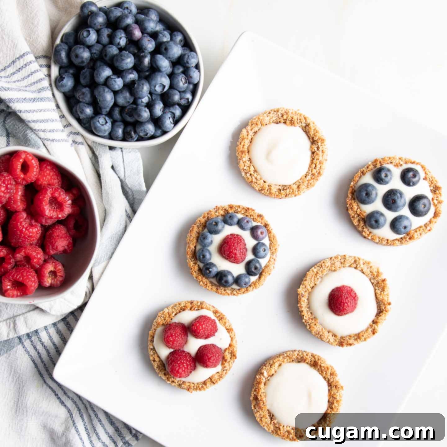 A vibrant display of fresh berries: a bowl of plump blueberries, a bowl of bright red raspberries, and a platter featuring an assortment of mini blueberry tarts. Some mini tarts are filled with the white tofu cream, while others are topped with a colorful mix of berries.
