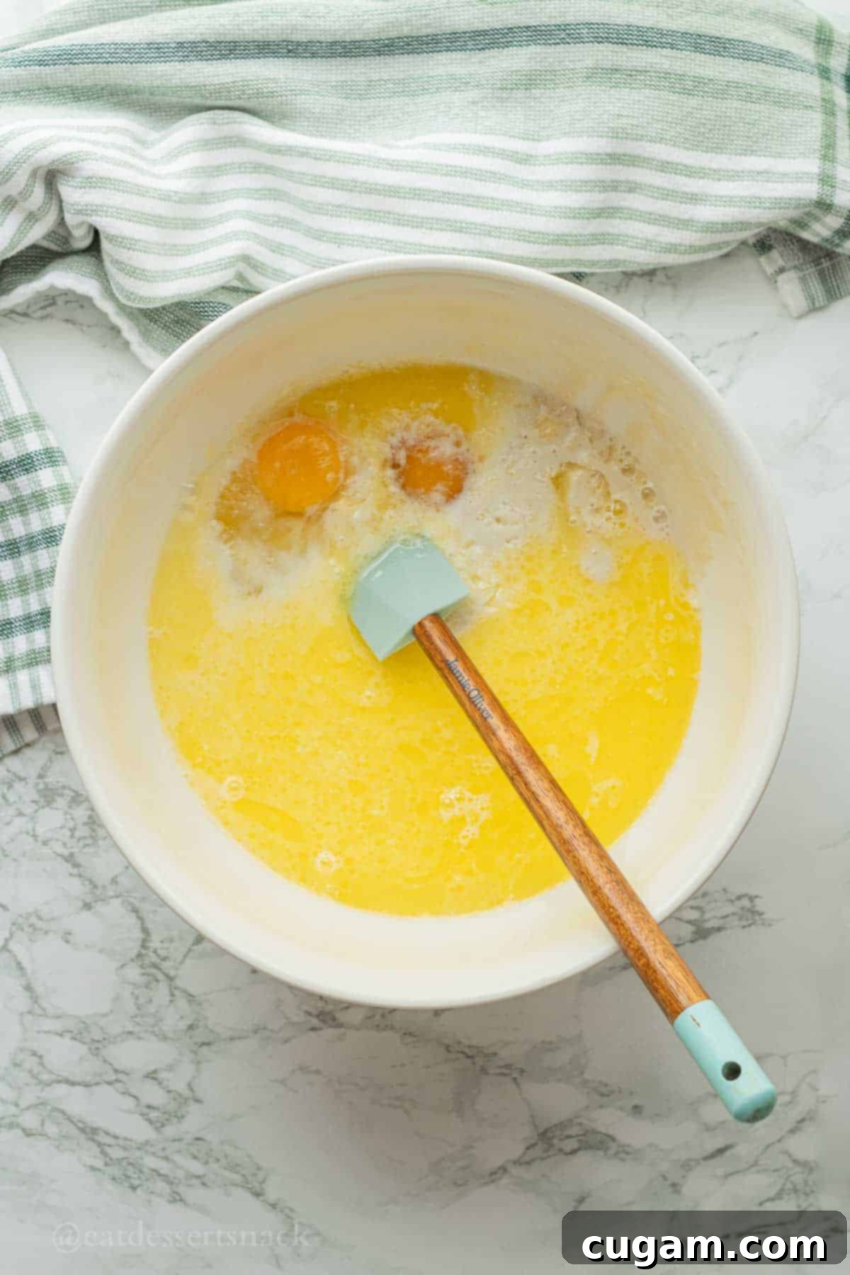 Eggs, melted butter, milk, and sourdough discard in white bowl with a rubber spatula, ready to be whisked.