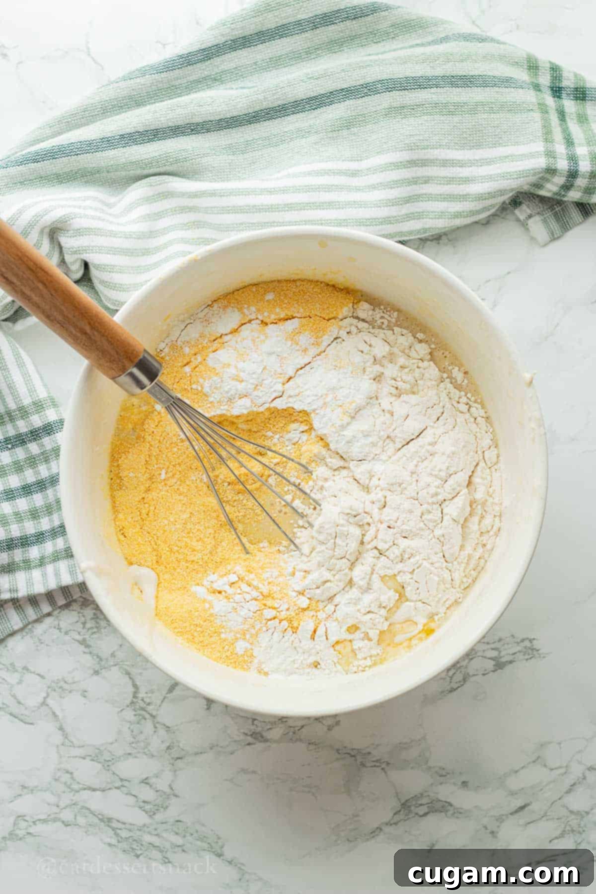 Cornmeal, flour, salt, and baking powder being sifted over the wet ingredients in a white bowl, next to a whisk.