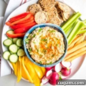 Blue bowl filled with Roasted Eggplant Dip sprinkled with parsley and paprika on top of a white plate with brightly colored veggies for dipping.
