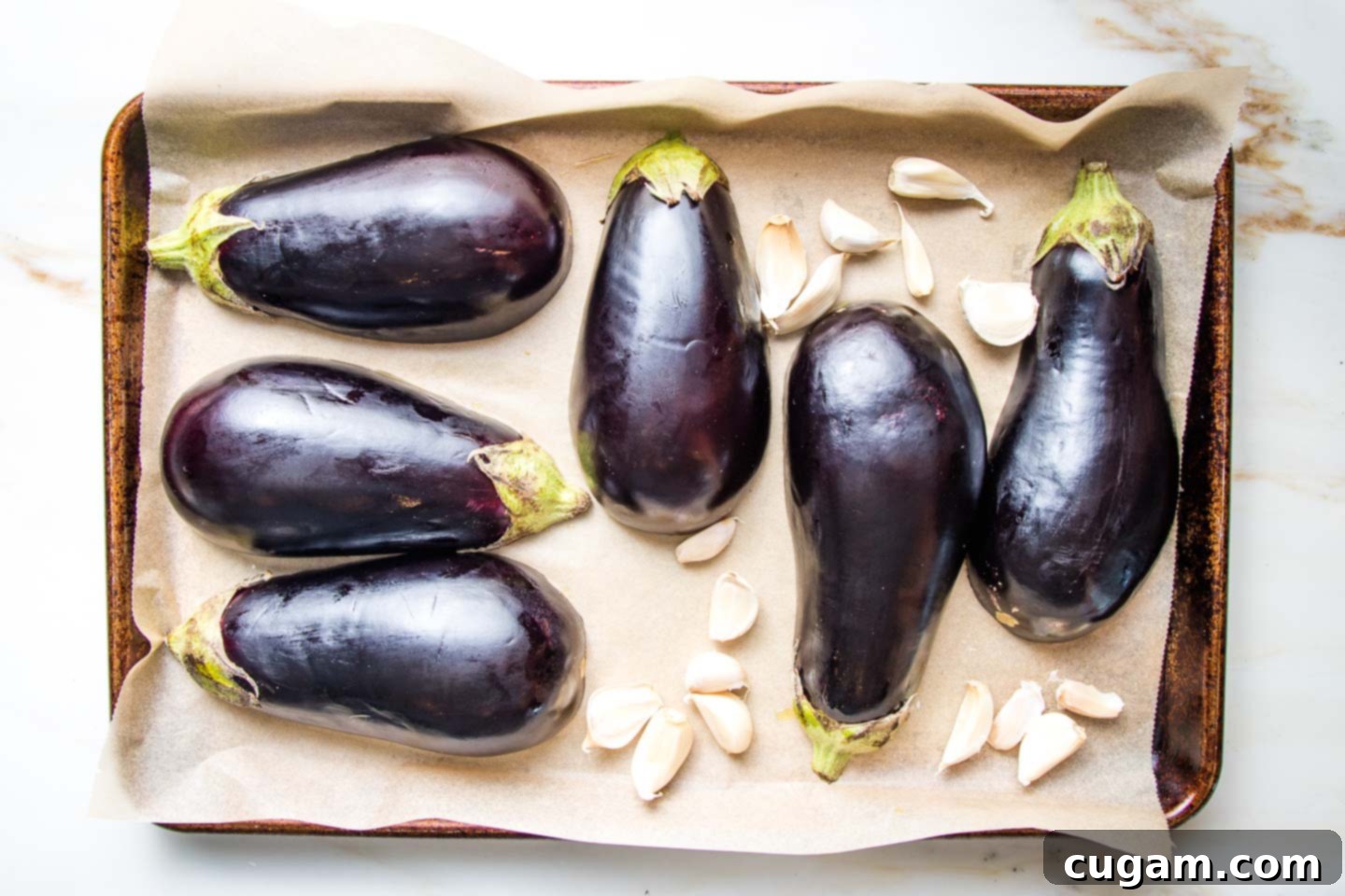 Halved eggplant and garlic cloves with their skins on a parchment lined baking tray.