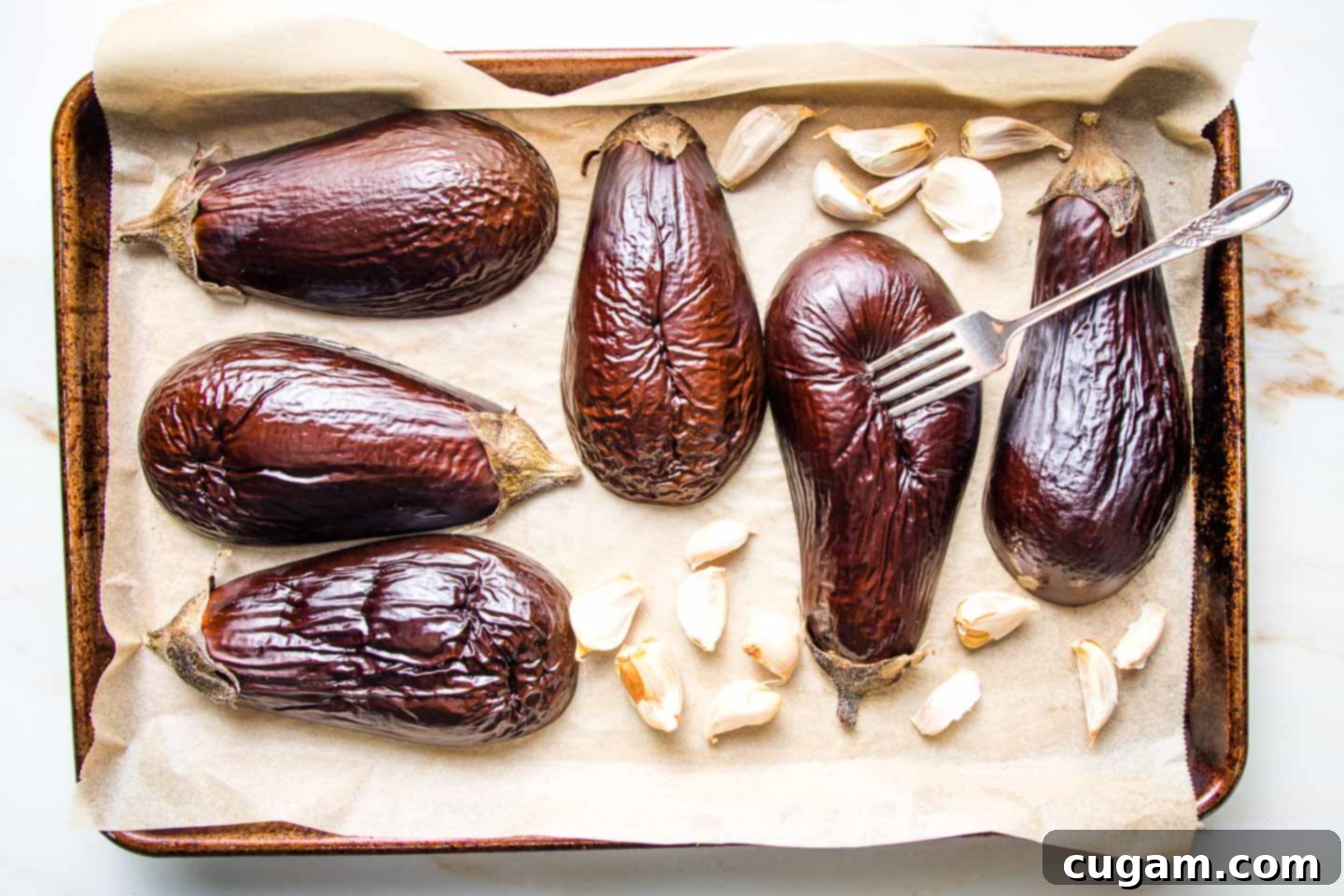 Halved eggplant roasting on a parchment lined baking tray with garlic cloves in their skins.