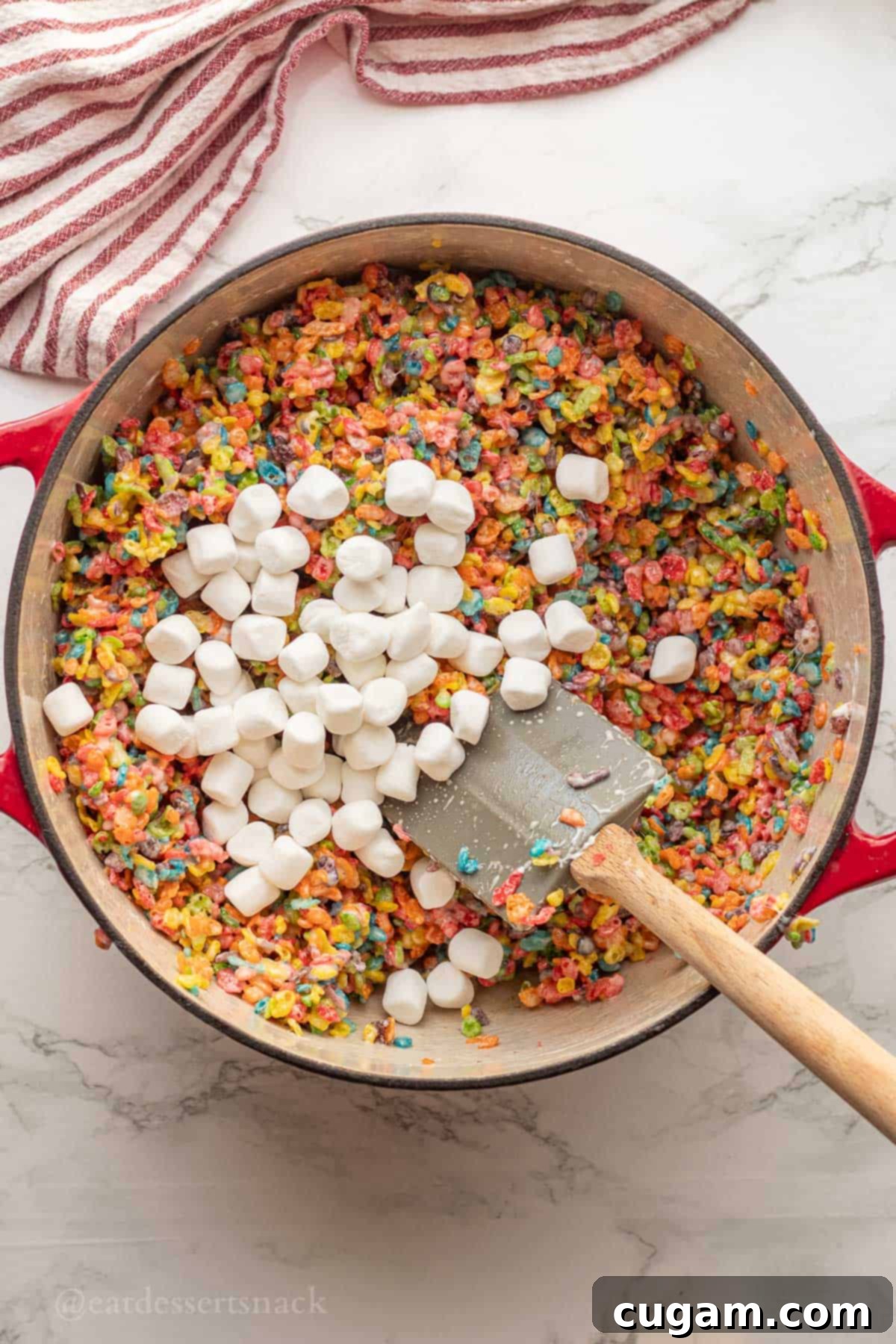 Reserved marshmallows being stirred into the Fruity Pebble and marshmallow mixture in a red pot with a rubber spatula.