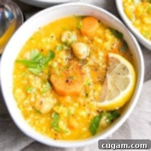 Nourishing Vegan Lemon Chickpea Rice Soup 15 White bowl filled with thick soup. Bright yellow broth with chickpeas, rice, carrots, lemon wedge and sprinkled with chopped parsley, salt and pepper.