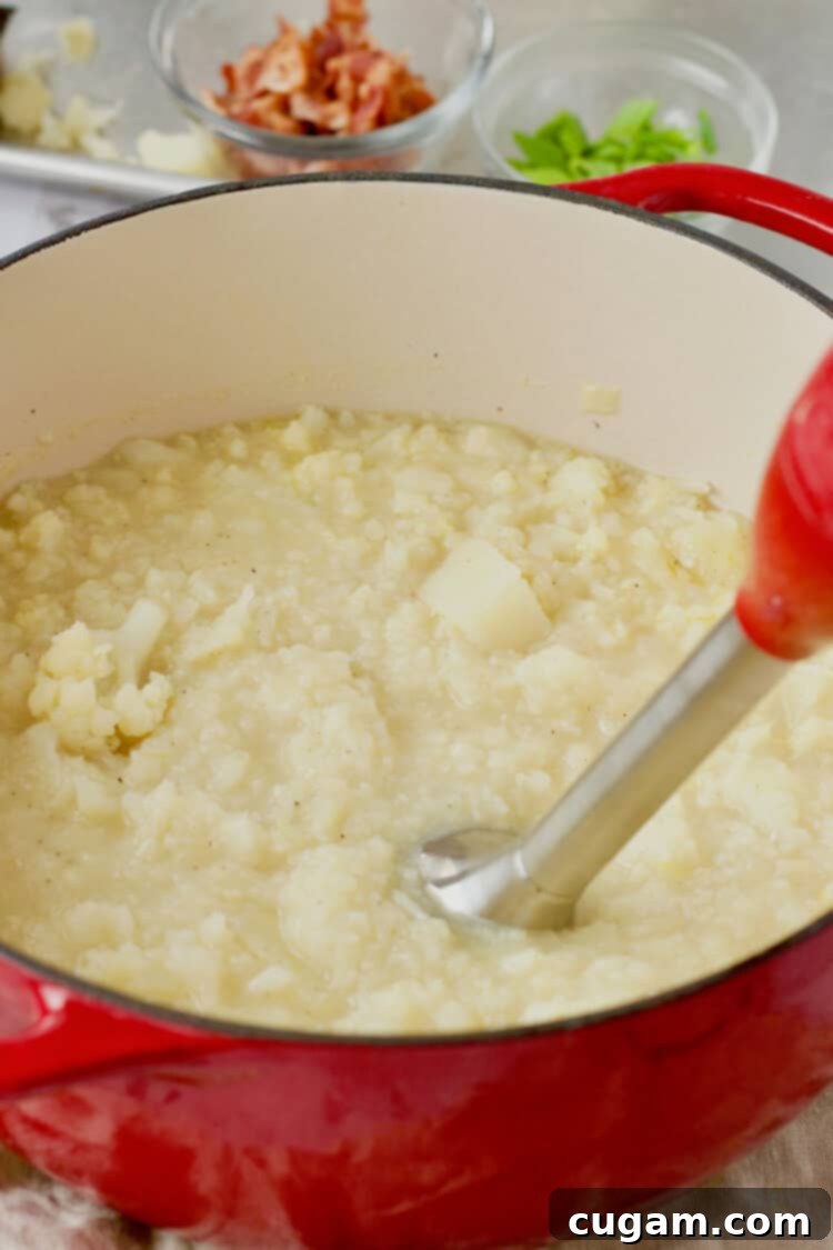 An immersion blender actively blending cooked cauliflower and potatoes into a chunky soup directly in a red pot.