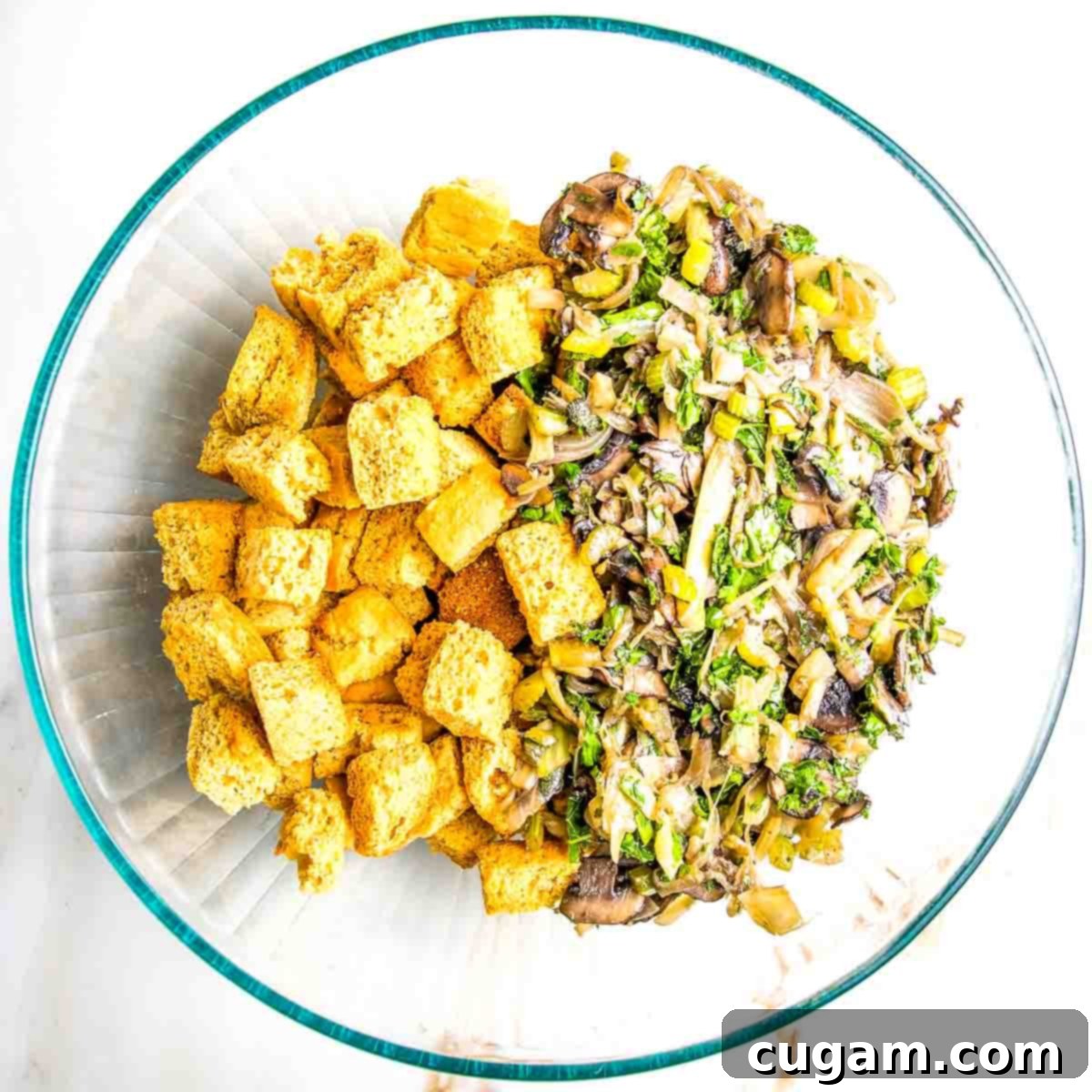 Glass mixing bowl containing dried cornbread croutons blended with sautéed vegetables, fresh herbs, and mushrooms, ready for the next step.