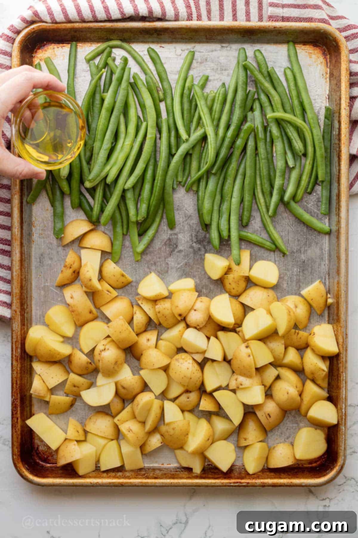 Pouring olive oil over raw green beans and potatoes on metal baking sheet.
