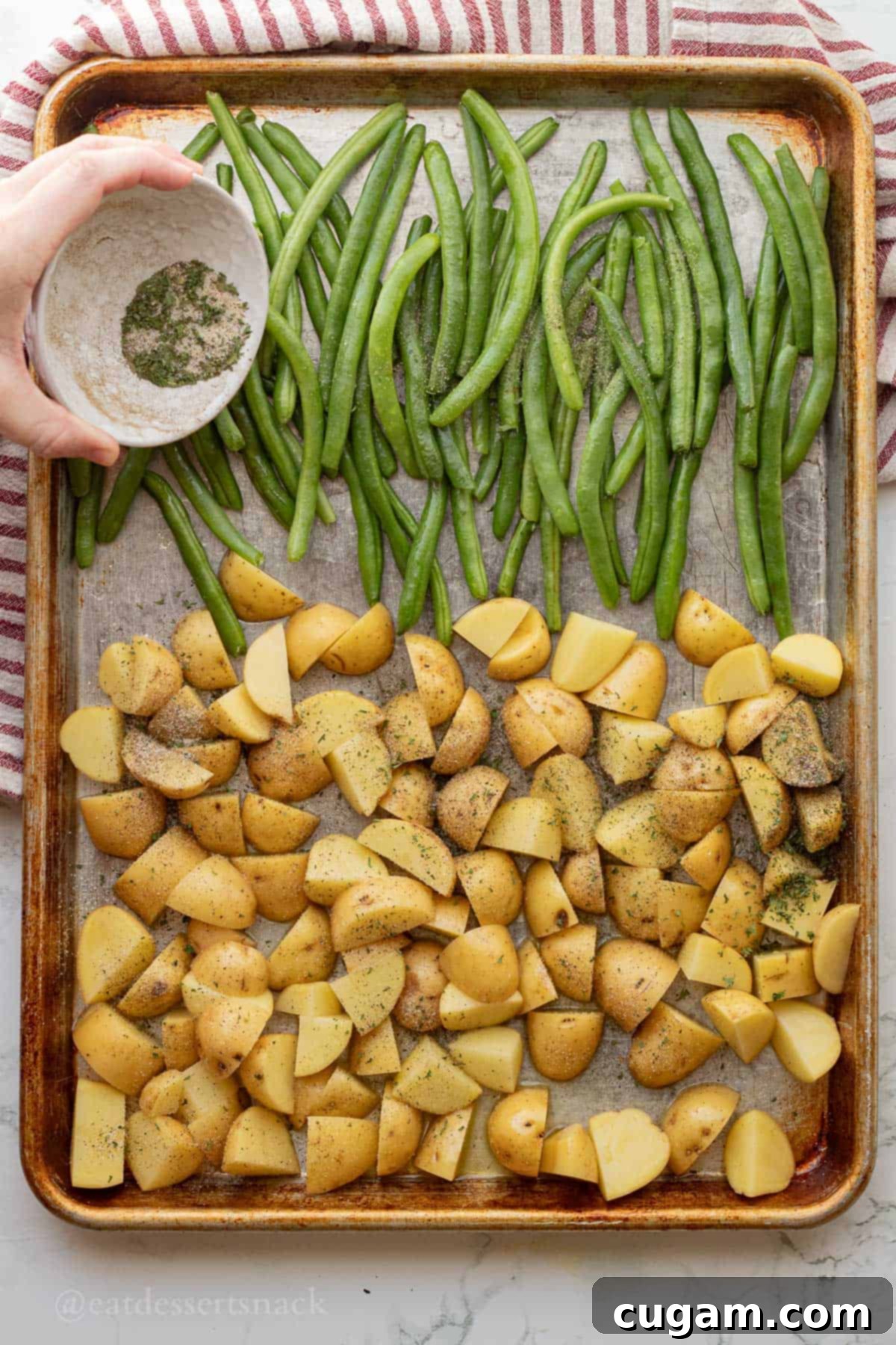 Sprinkling spices on metal pan of raw green beans and potatoes.