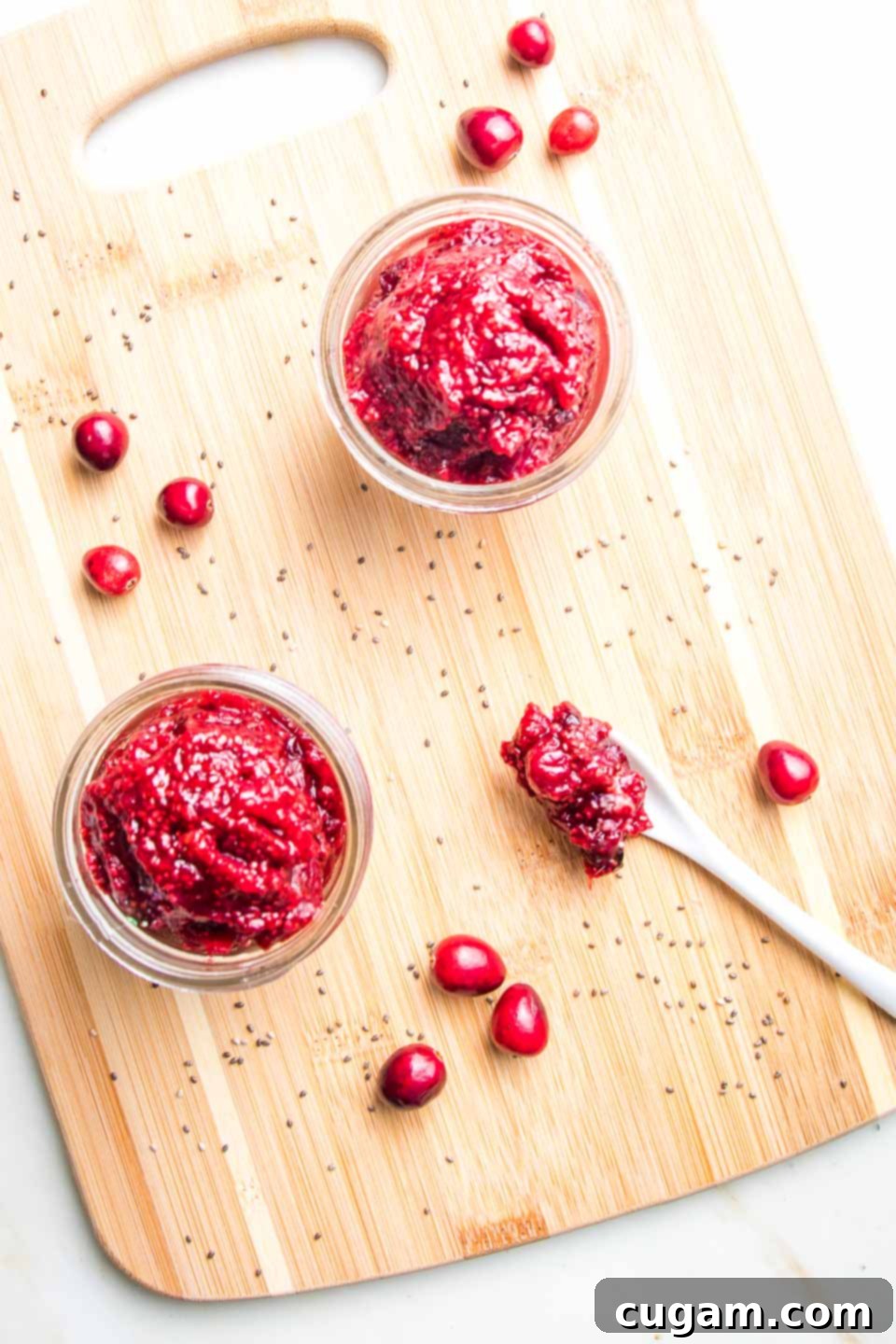 Small jar of cranberry chia jam on a wooden board with a few fresh cranberries and some chia seeds sprinkled about.
