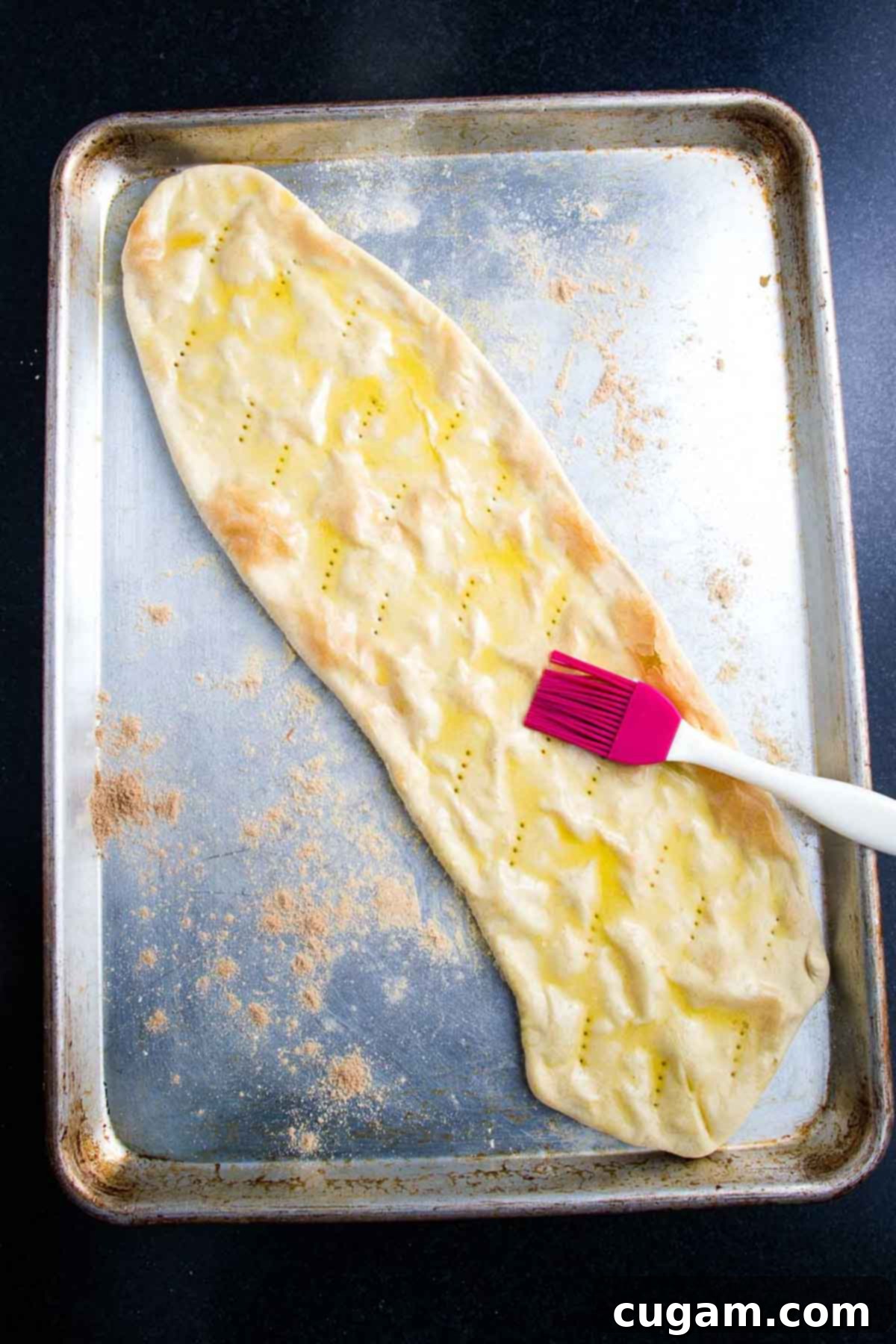 A flatbread crust on a baking tray is being brushed with olive oil using a pastry brush.