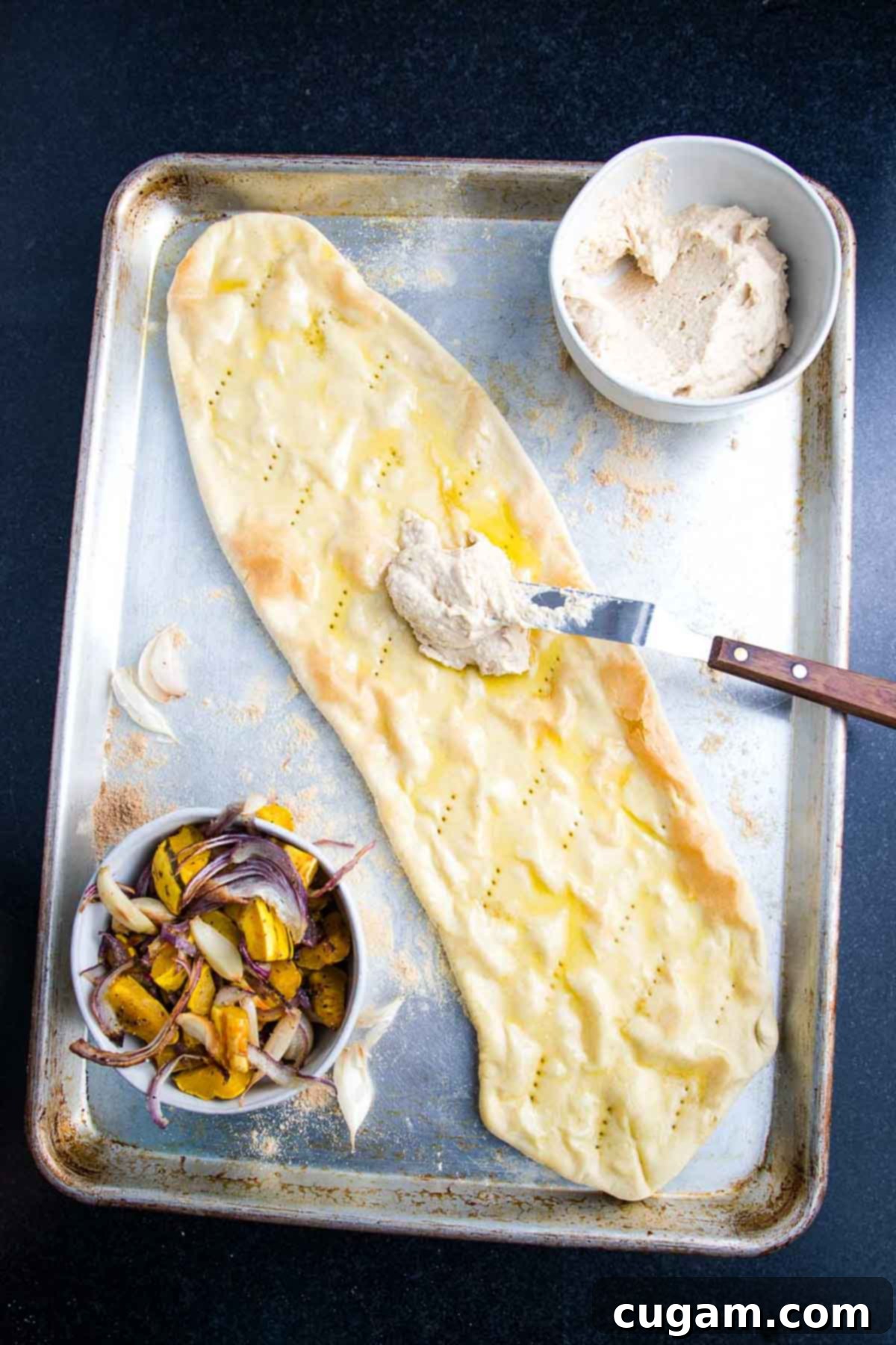 Hummus being spread evenly onto an olive oil-brushed flatbread pizza crust.