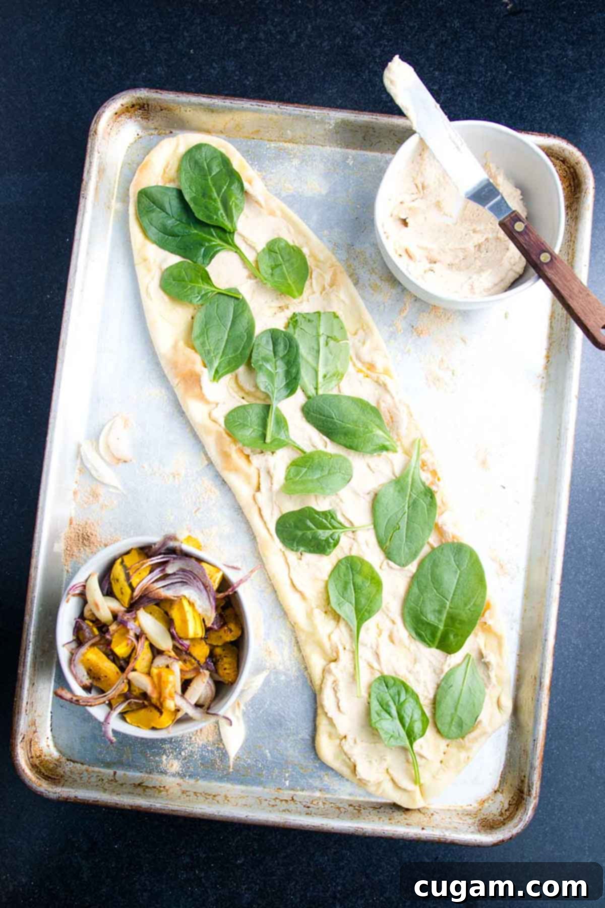 A flatbread pizza crust, spread with hummus, is being topped with fresh baby spinach leaves.