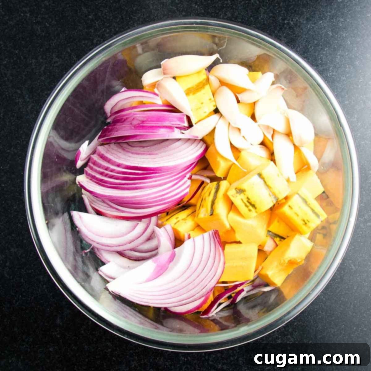 A mixing bowl filled with cubed delicata squash, thinly sliced red onions, and whole unpeeled garlic cloves.