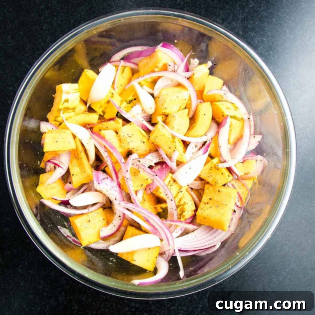Cubed squash, sliced onions, and garlic cloves are generously drizzled with olive oil and seasoned with salt and pepper in a bowl.