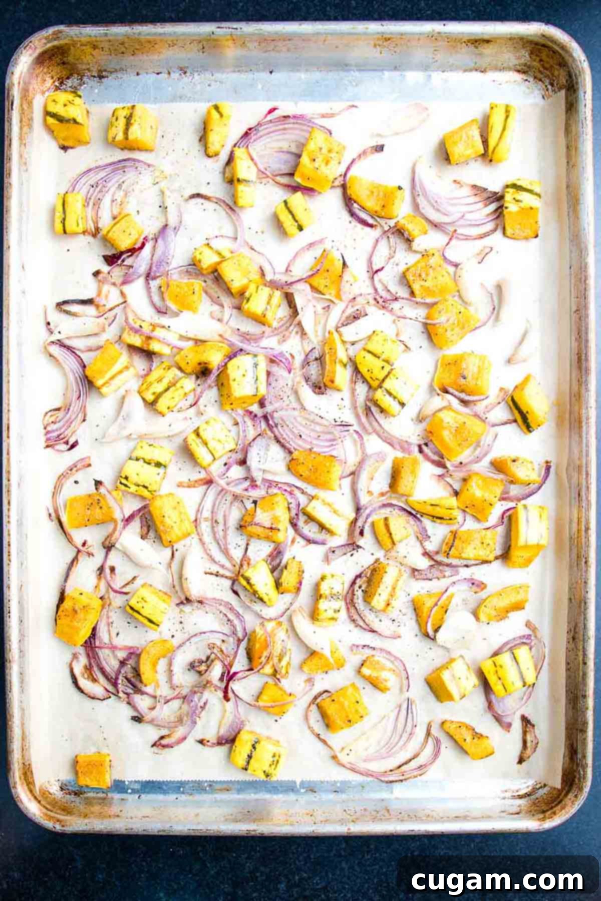 A baking sheet is filled with a single layer of roasted cubes of squash, thinly sliced onions, and whole cloves of garlic, ready for the oven.