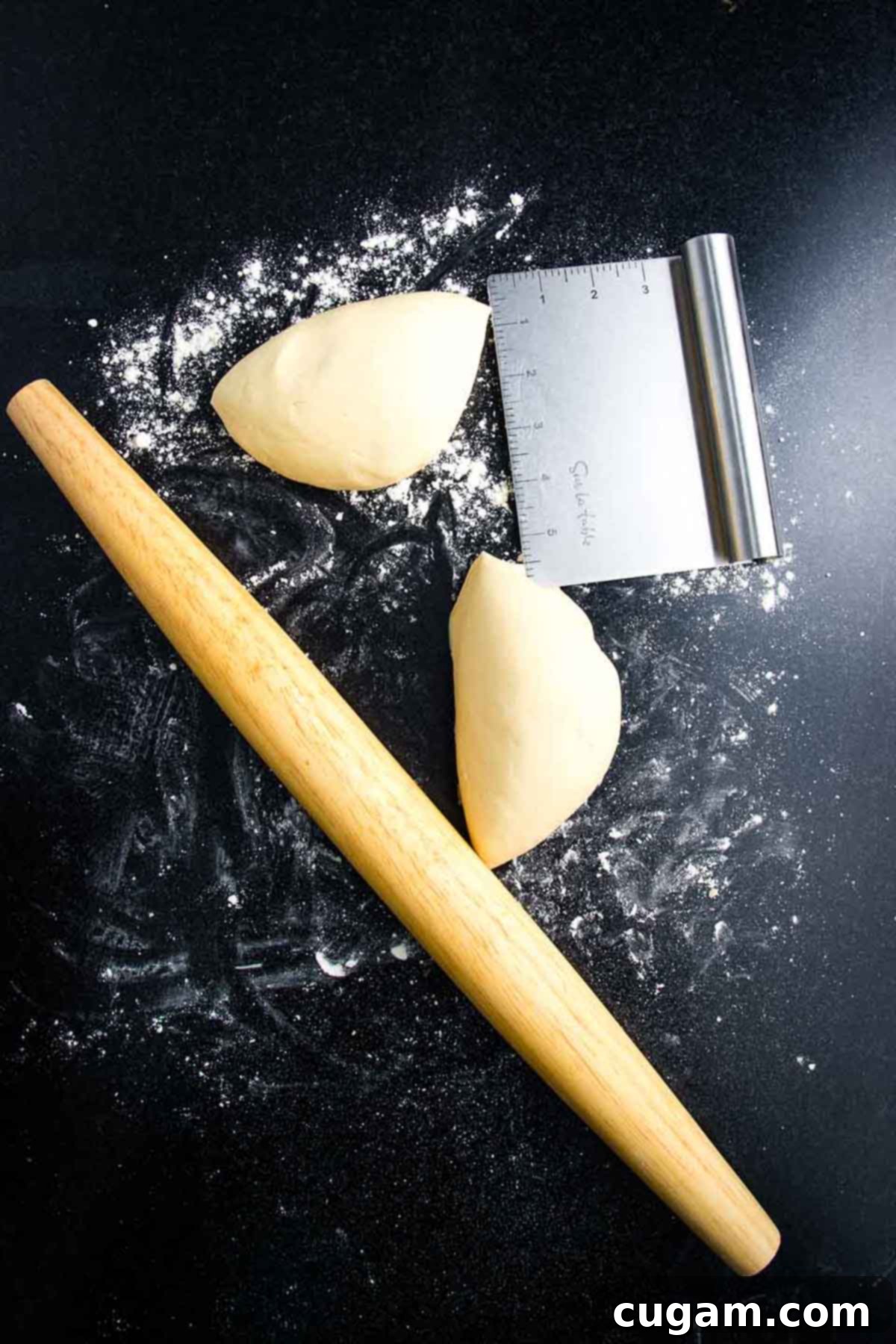 A ball of pizza dough, cut in half, rests on a floured black surface with a wooden rolling pin nearby.