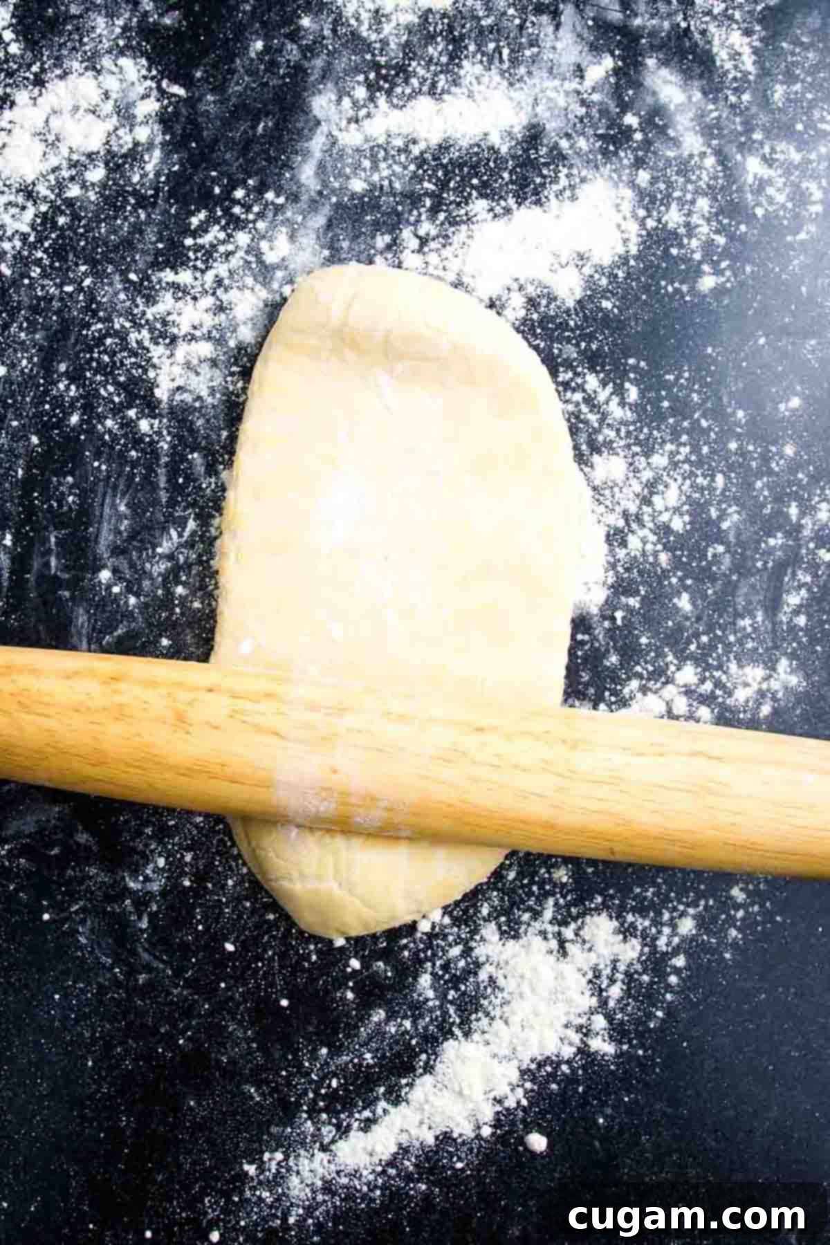 A wooden rolling pin is used to roll out a portion of pizza dough on a floured black surface.
