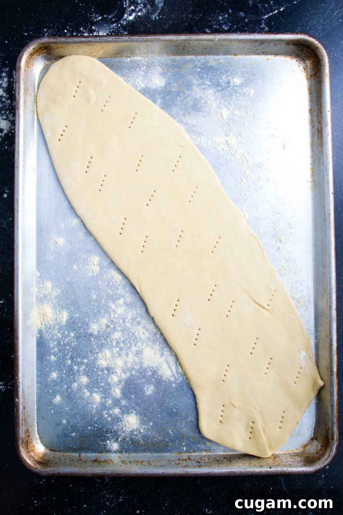 A long, thin oval of pizza dough on a baking sheet, marked with fork pricks across its surface.