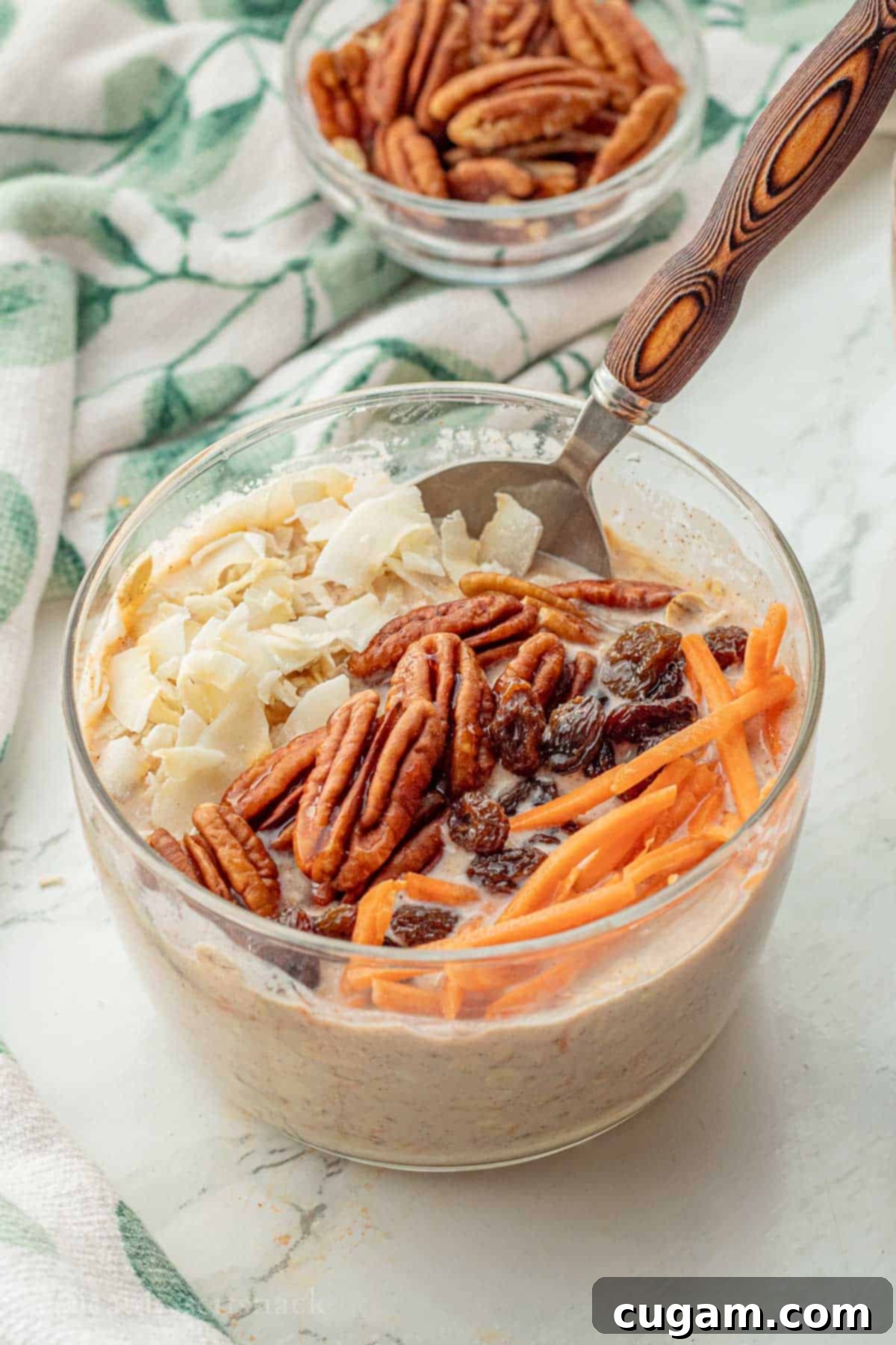 Pecans, raisins, carrots, and coconut on bowl of carrot cake overnight oats with spoon.