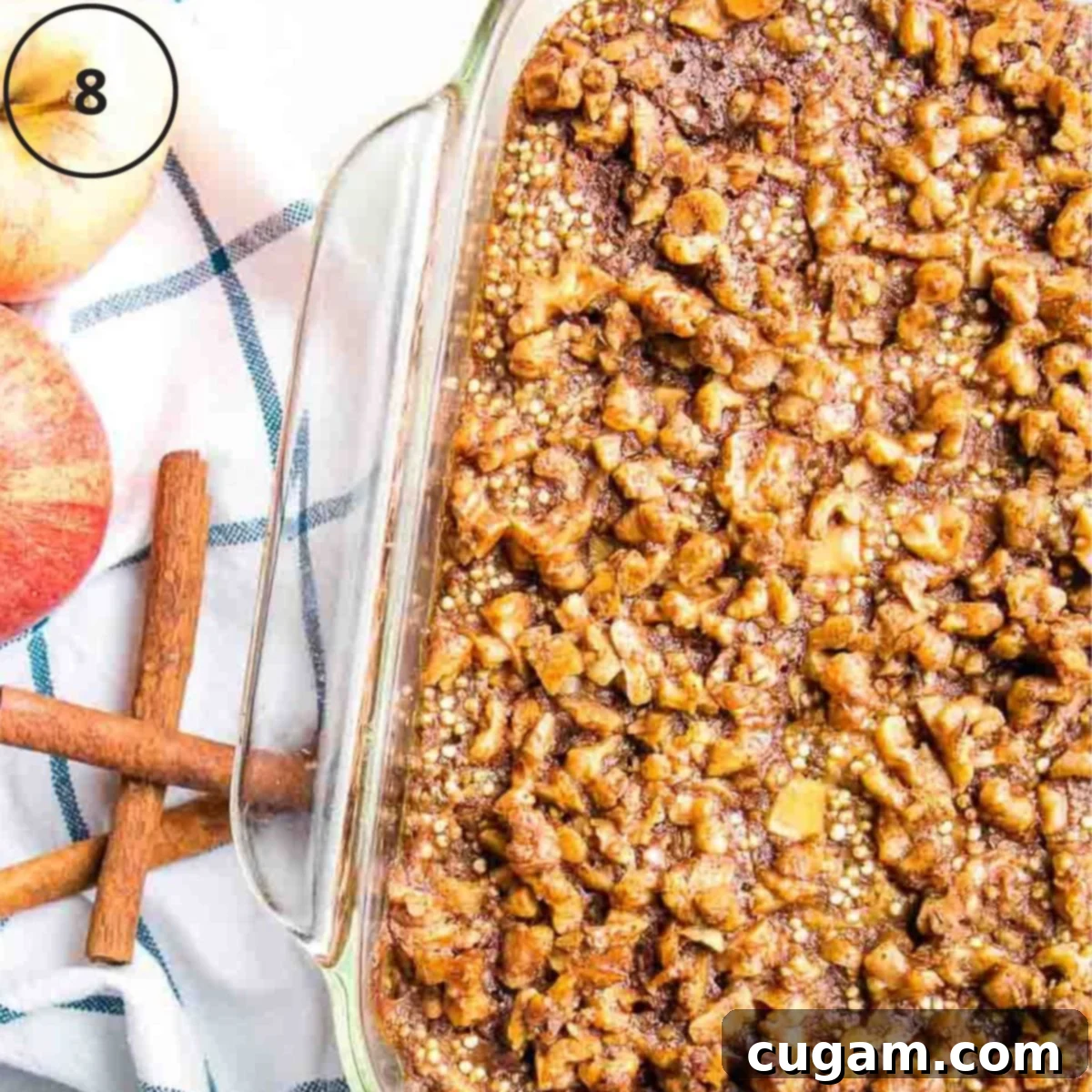 A square glass baking pan filled with freshly baked, golden-brown quinoa. In the background, there are fresh apples and elegant cinnamon sticks, hinting at the warm, inviting flavors within the dish.