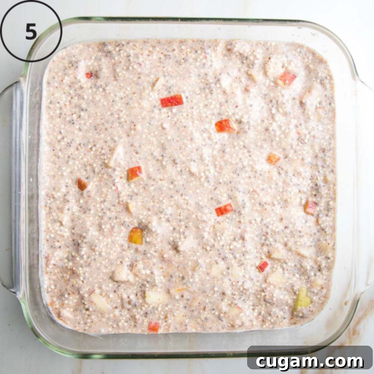 A square glass baking dish neatly filled with the rich quinoa batter, dotted generously with vibrant diced red apples, skin still on, presenting a beautiful base for the breakfast bake.