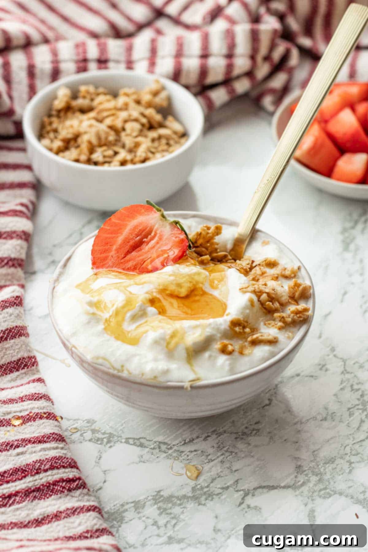 White bowl of yogurt with honey, granola, and strawberry on top with gold spoon.