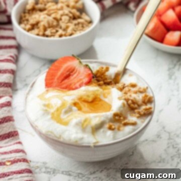 White bowl of yogurt with honey, granola, and strawberry on top with gold spoon.
