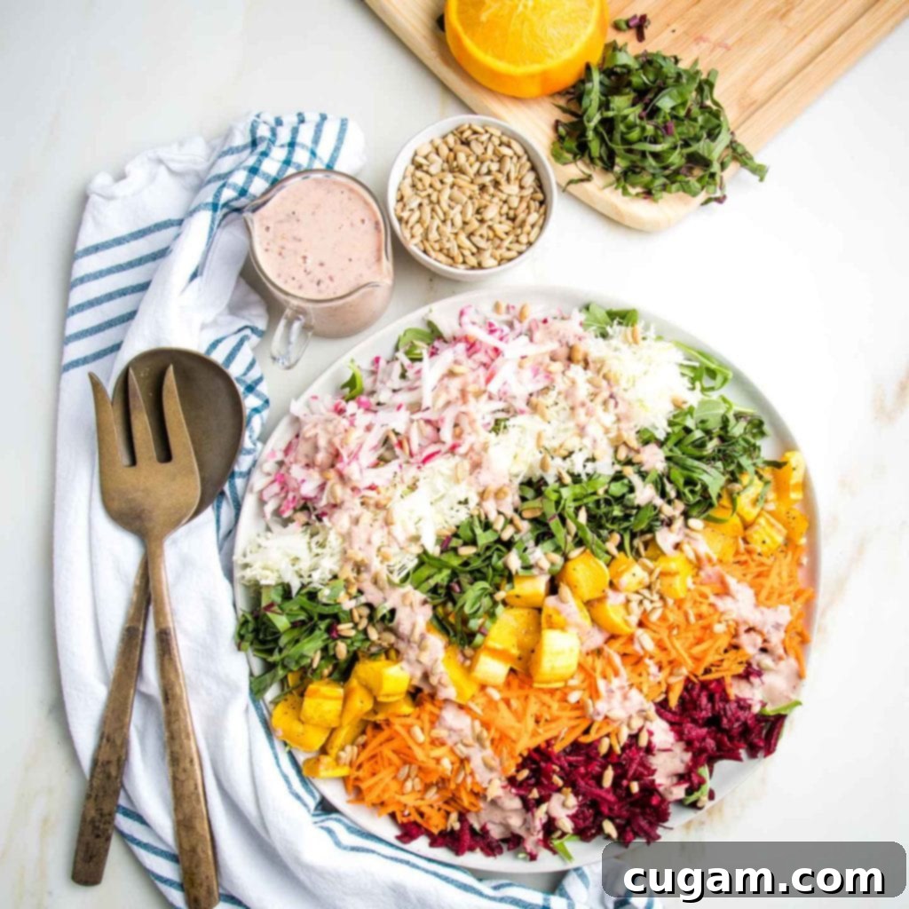 Rainbow salad on a white plate with a pink dressing drizzled across the colorful stripes. Serving pieces on a white tea towel with blue stripes, a small pitcher of extra dressing plus some sunflower seeds and a background cutting board with julienned beet greens and some oranges that have already been squeezed.