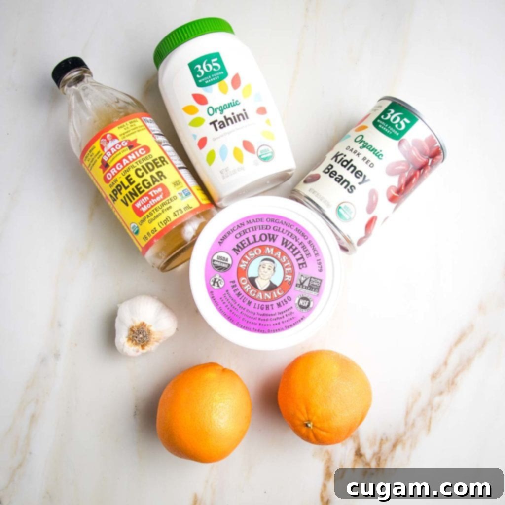 Salad Dressing Ingredients on a marble counter: bottle of apple cider vinegar, head of garlic, 2 oranges, jar of tahini, can of red kidney beans, container of light miso paste.