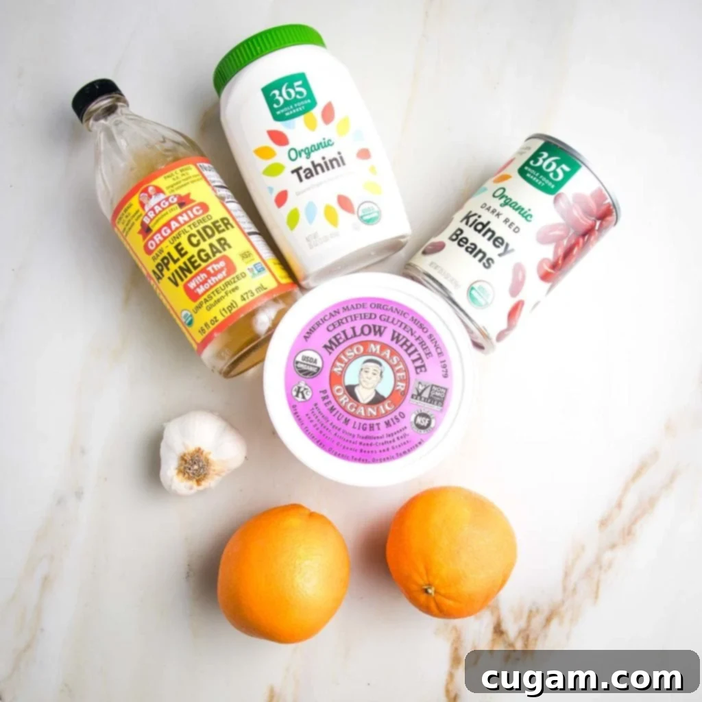 Kaleidoscope Nourish Bowl 4 Salad Dressing Ingredients on a marble counter: bottle of apple cider vinegar, head of garlic, 2 oranges, jar of tahini, can of red kidney beans, container of light miso paste.