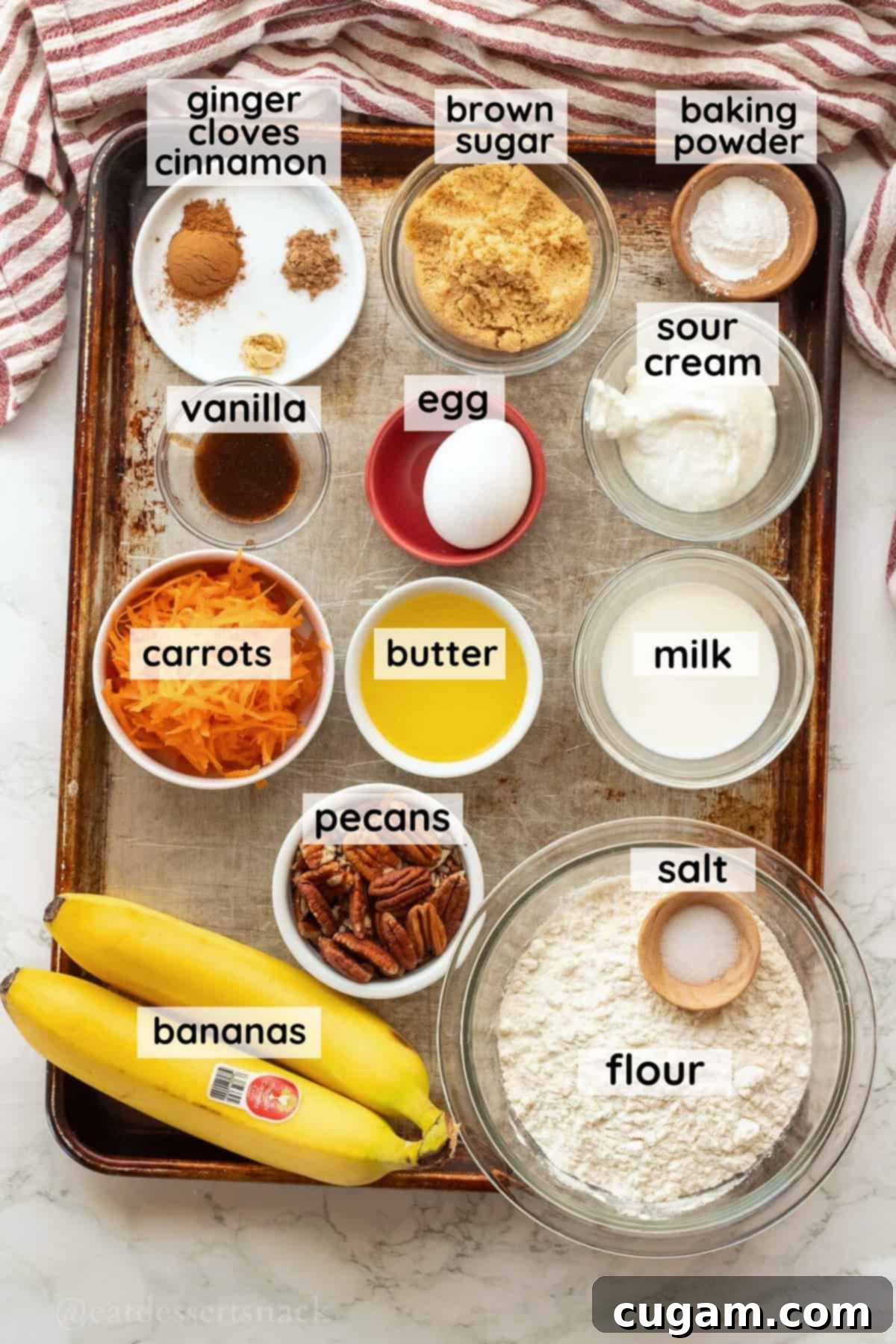 Labeled ingredients for banana carrot muffins in glass bowls on metal baking pan.