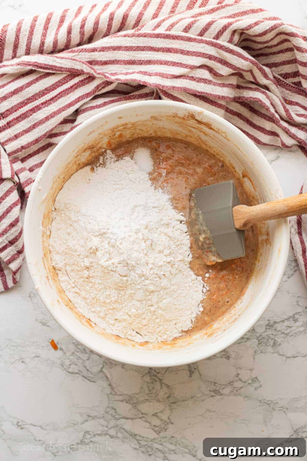Flour, salt, baking powder in bowl of carrot banana muffin batter with rubber spatula.