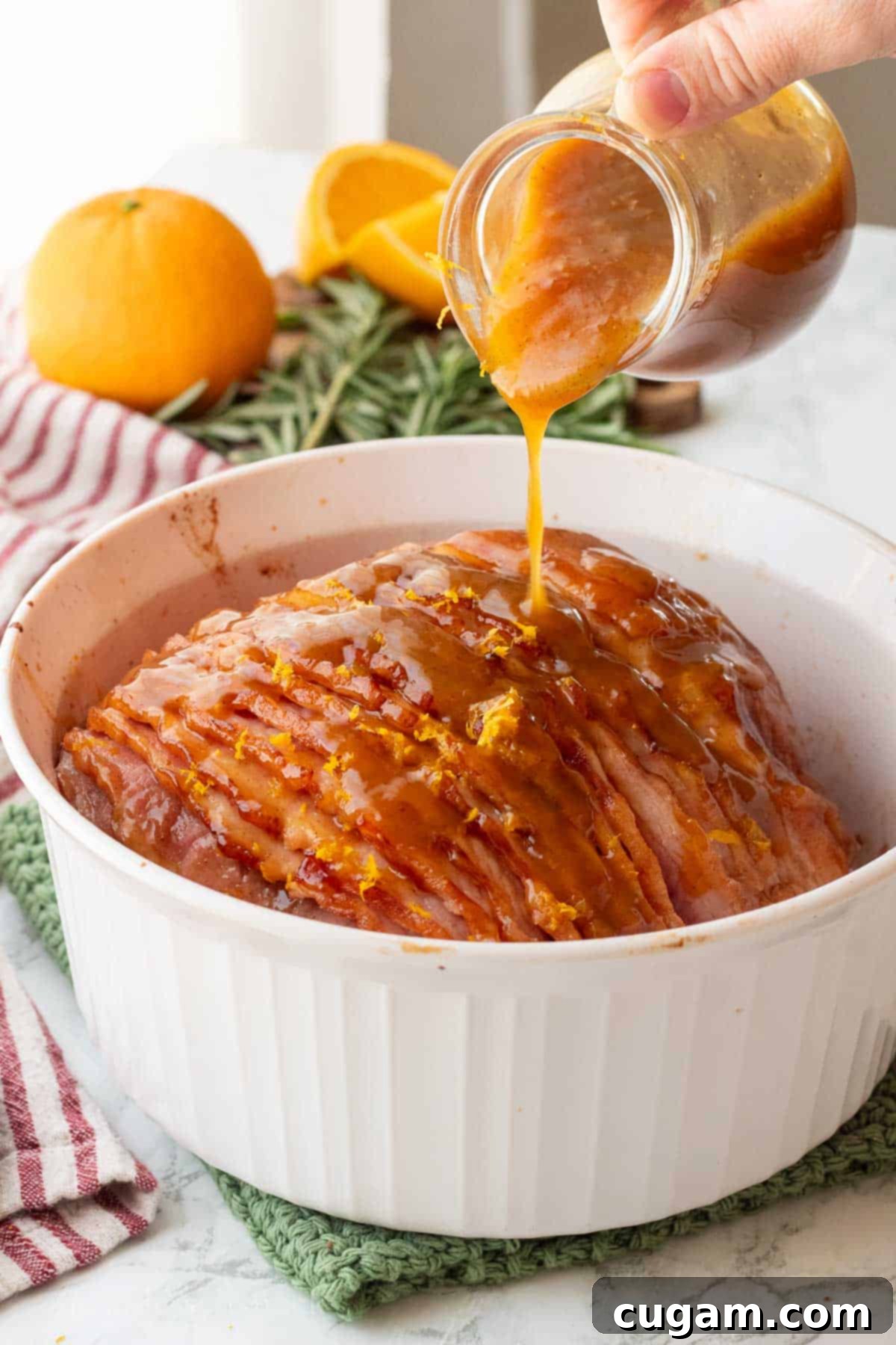 A hand pouring glossy orange glaze over a cooked spiral ham in a glass baking dish, ensuring every crevice is coated.
