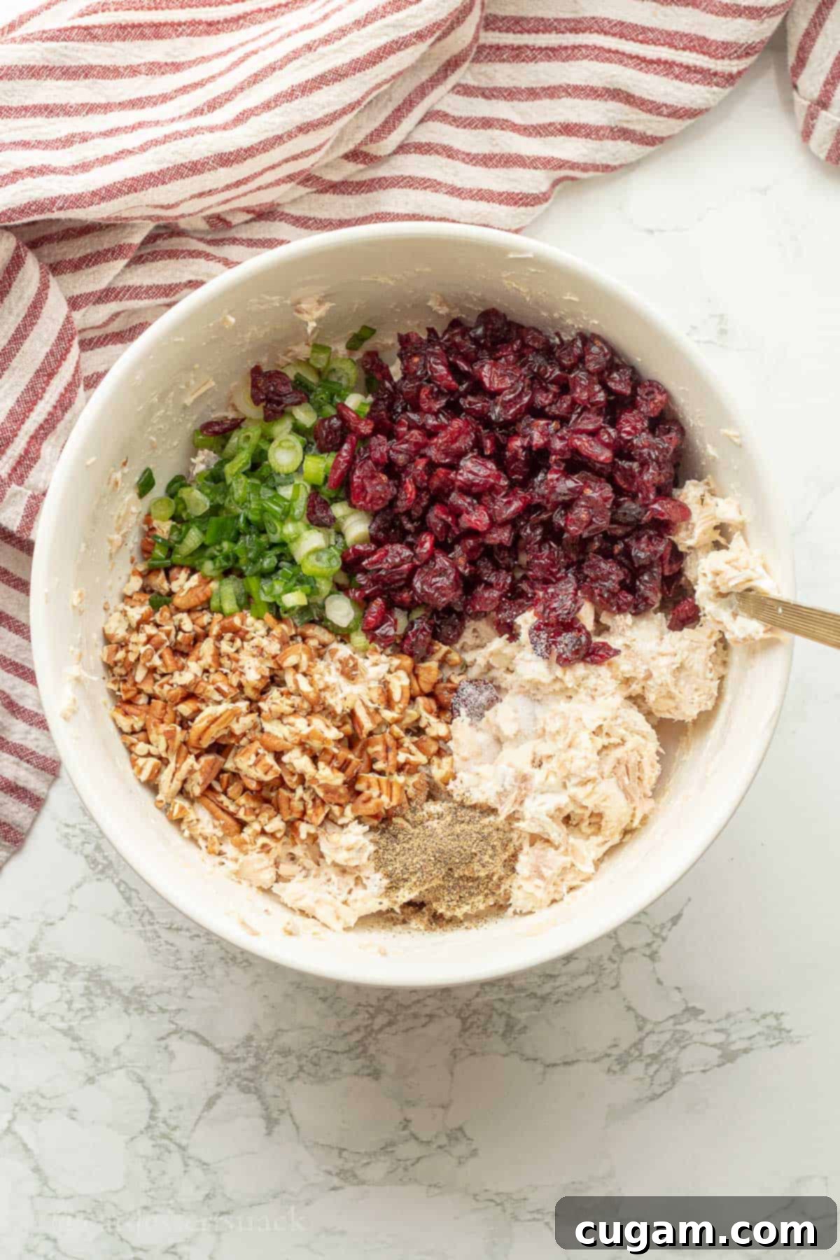 All ingredients for chicken cranberry salad in a white bowl, being mixed with a gold spoon.