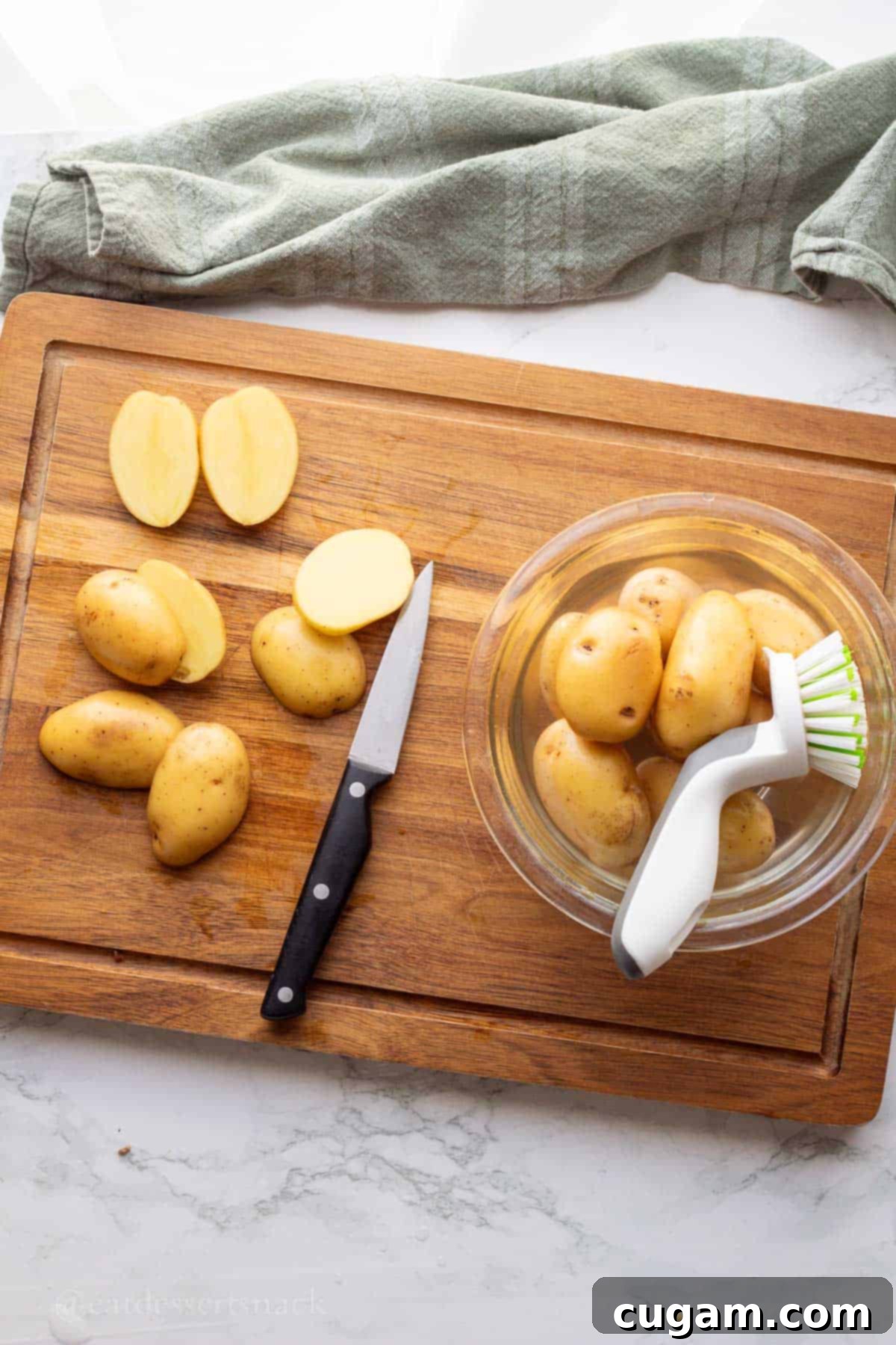 Bowl of water with raw baby potatoes and scrub brush on wood cutting board with knife and sliced potatoes.