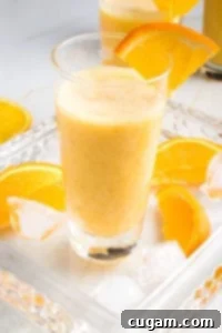 Creamy Orange Bliss 11 Glass filled with a frothy Orange Smoothie, with a wedge of orange on the side of the glass, more orange wedges on the glass serving tray along with some ice cubes.