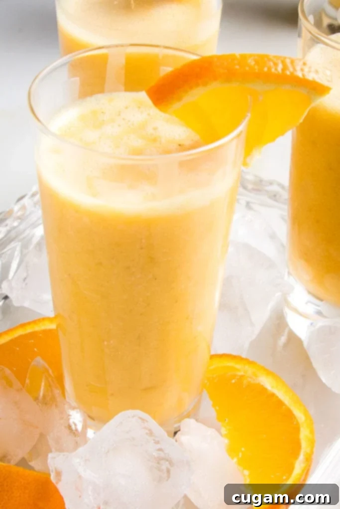 Creamy Orange Bliss 3 Close-up of a glass filled with a frothy orange smoothie, garnished with a fresh orange slice, sitting on a serving tray with scattered ice cubes and orange slices.