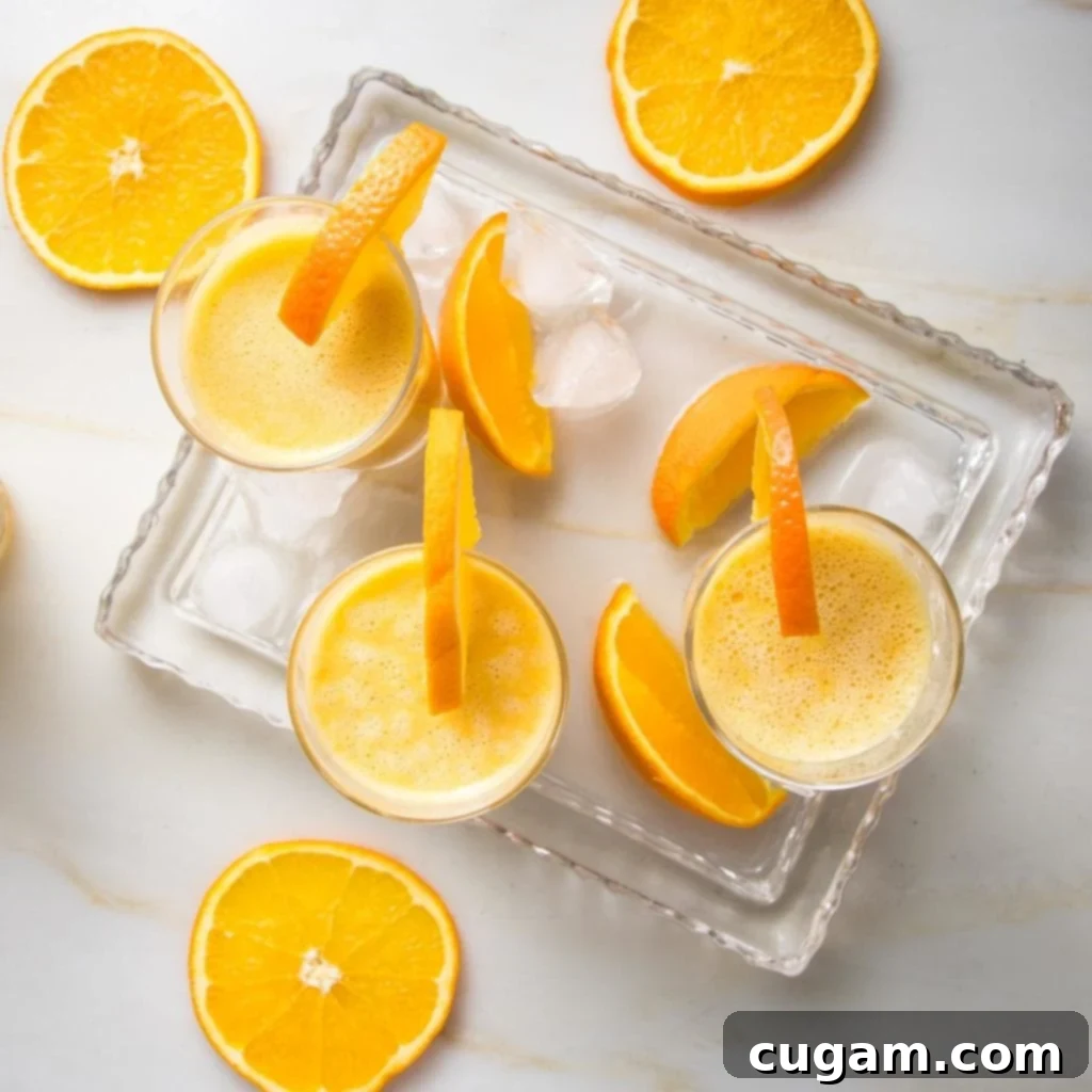 Creamy Orange Bliss 9 Three Orange Julius smoothie glasses, garnished with orange wedges, resting on a glass tray. Ice cubes and orange slices are scattered around, creating a fresh and inviting scene.