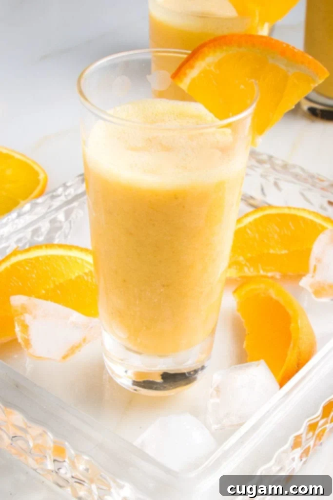 Creamy Orange Bliss 10 A single glass of frothy Orange Julius Smoothie, perfectly blended and garnished with an orange wedge, resting on a serving tray surrounded by ice cubes and extra orange slices.