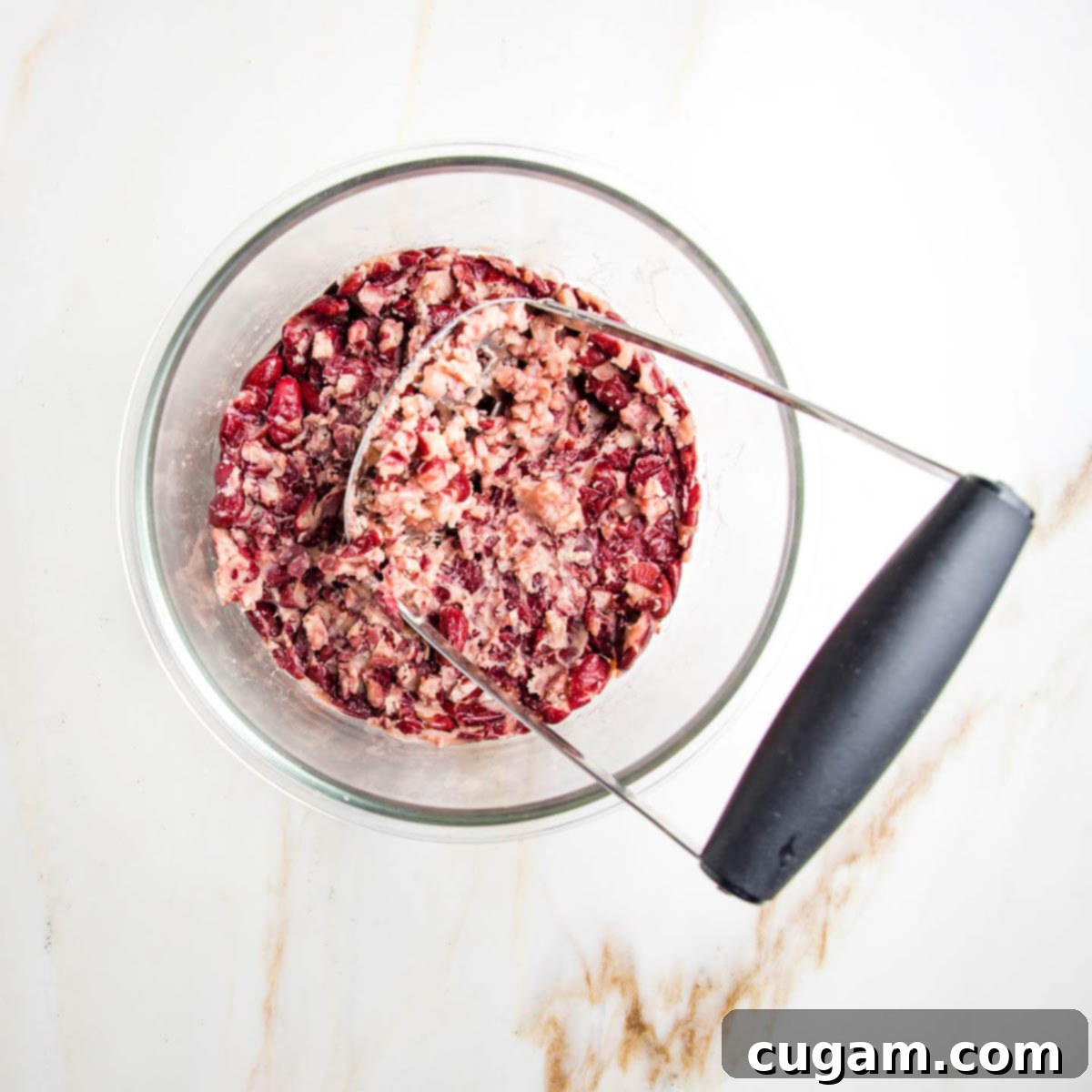Potato masher squishing red kidney beans in a glass bowl to create a paste-like texture.