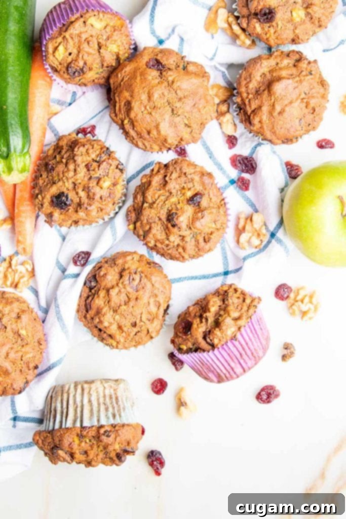 Morning Glory Muffins, tumbled out onto a white dish towel with blue pattern, along with a zucchini, two carrots and a green apple. There are walnuts and dried cranberries strewn around. 