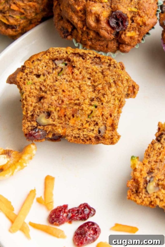 Breakfast muffin that has been cut in half. You can see pieces of shredded carrots and zucchini plus walnuts and dried cranberries. There are a few cranberries and shredded carrots in the foreground on a white plate.