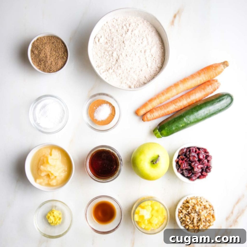 Various muffin ingredients laid out on a white surface, including shredded carrots, zucchini, apple, dried cranberries, walnuts, flour, and spices.