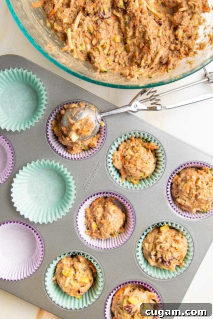 Bowl of muffin batter and a muffin tin lined with purple and light blue liners, half of them already filled with batter and a large cookie scoop filling another. 