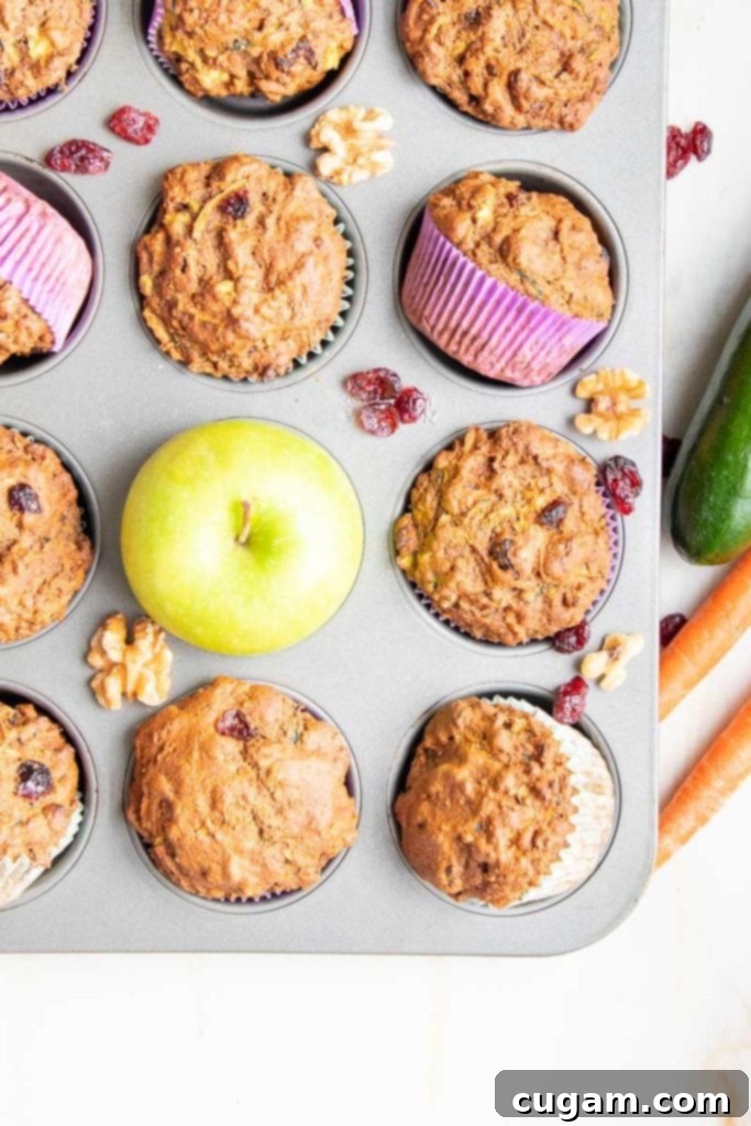 Muffin tin filled with morning glory muffins, one has a green apple, there are dried cranberries and walnuts strewn about and zucchini and carrots off to the side