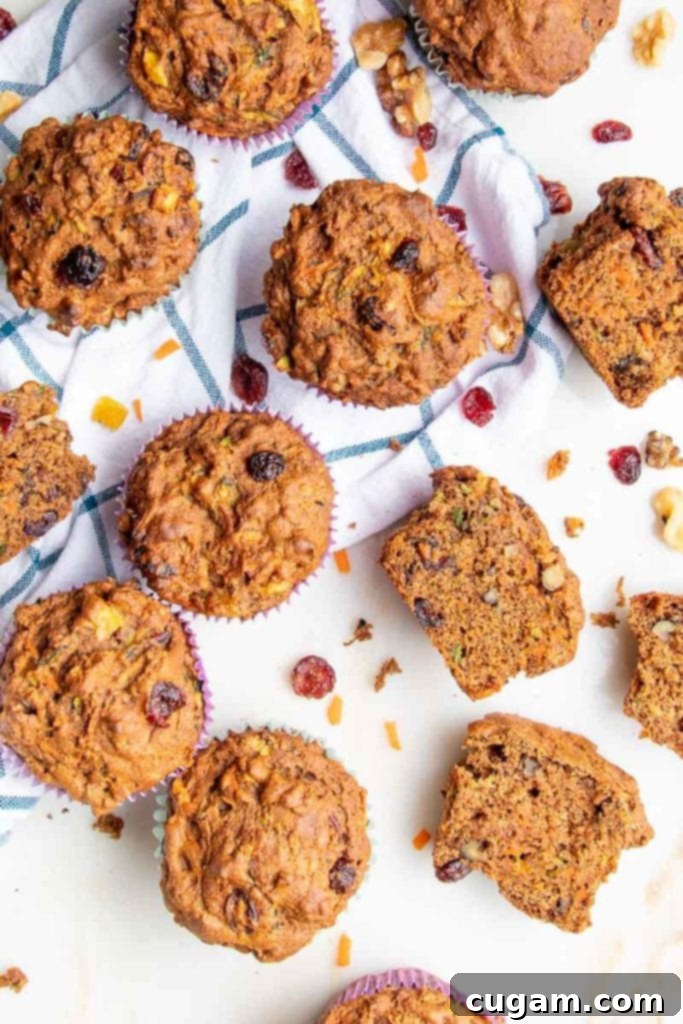 A bunch of muffins scattered on a tea towel with blue wide plaid. some muffins are cut open to see the insides, with walnuts, shredded carrots and zucchini plus dried cranberries. 